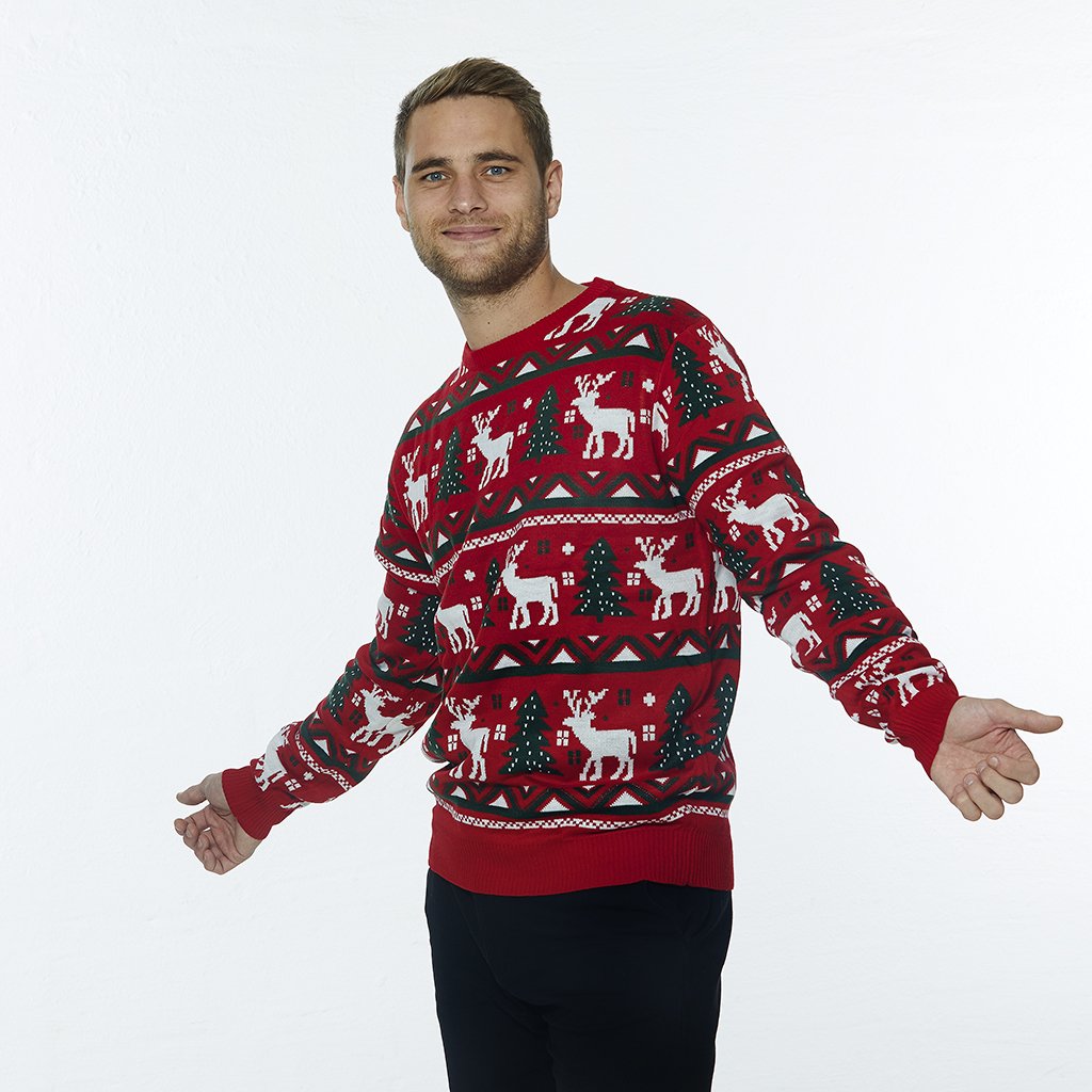 Een poserende man die een rode kersttrui draagt met rendieren en kerstbomen erop.